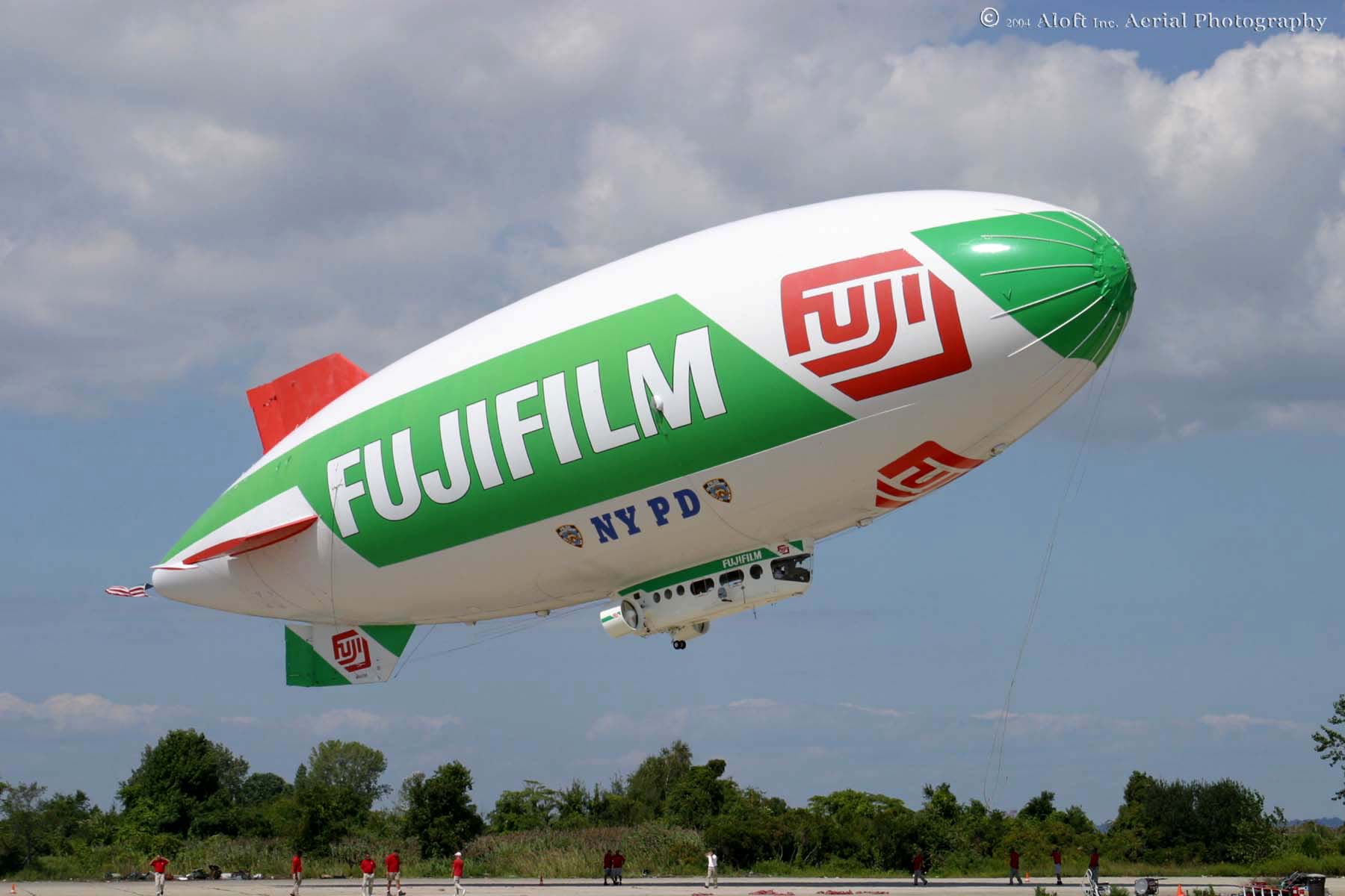 A large white and green blimp flying over the grass.