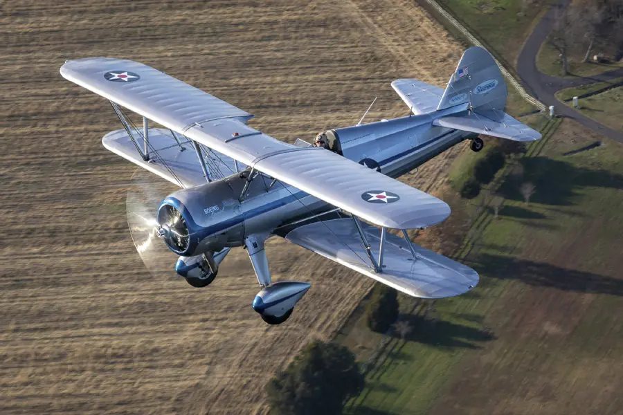 A silver biplane flying over an open field.